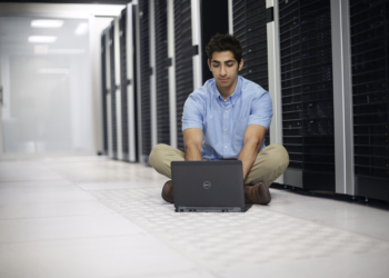 Young South Asian man sitting on the floor in a large data center working on a Dell Latitude 12 7000 Series (Model E7240) Touch notebook computer, in front of a row of server racks with Dell enterprise products.