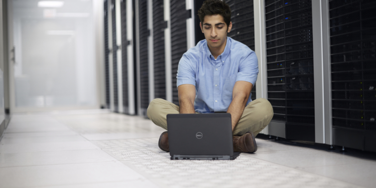 Young South Asian man sitting on the floor in a large data center working on a Dell Latitude 12 7000 Series (Model E7240) Touch notebook computer, in front of a row of server racks with Dell enterprise products.