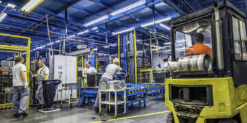 Factory workers doing their jobs while a forklift is driving by.