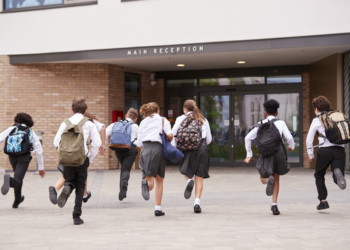 Group Of High School Students Wearing Uniform Running Into School Building At Beginning Of Class